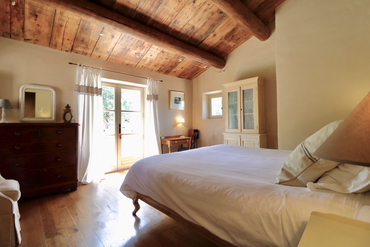 A bright and cozy bedroom featuring a large bed with crisp white bedding, wooden ceiling beams, an antique dresser, a desk by the window, and a door leading to a sunny outdoor area.